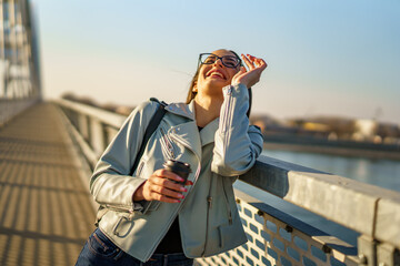 Happy beautiful young woman enjoying in sunset, she's walking on city bridge while holding phone...