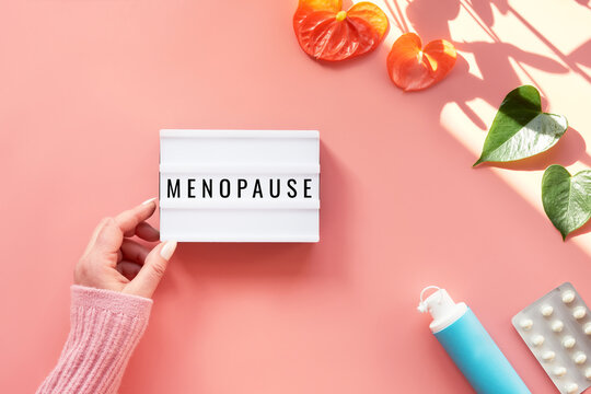 Text Menopause on light box in hand. HRT Replacement hormone therapy, HRT concept. Pink background with exotic leaves, flowers, pills, estrogene gel. Sunlight, long shadows.