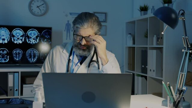 Tired doctor in white coat examining MRI lungs images taking off glasses feeling exhausted at night in dark office. Medicine and healthcare concept.