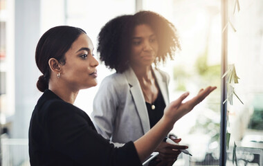 Adding structure to their ideas. Shot of two businesswomen brainstorming with notes on a glass wall...