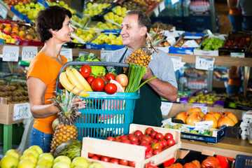 Smiling cheerful positive couple sellers is standing with basket with fruits and vegetables in the supermarket.