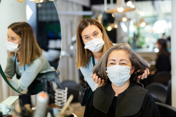 Elderly woman wearing protective face mask sitting in chair in hair salon while professional stylist proposing new hairstyle. New life reality during COVID 19 pandemic