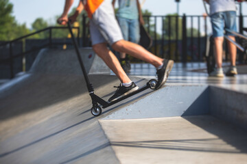 Fototapeta premium Skate park view with kids on a kick scooter doing tricks and stunts, boys in a skate park riding bmx bike and skate, playing and enjoying summer on new ramp, teenagers skateboarders and bmx riders