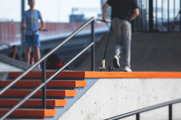 Fototapeta premium Skate park view with kids on a kick scooter doing tricks and stunts, boys in a skate park riding bmx bike and skate, playing and enjoying summer on new ramp, teenagers skateboarders and bmx riders
