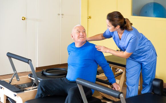 Young woman physiatrist helping aged man doing exercises on reformer as part of injury rehabilitation. Wellness concept for elderly
