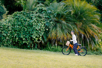Cute teen girl with glasses in a white long-sleeve shirt strolls an orange bicycle on the park lawn, smiling happily and looking at the camera.