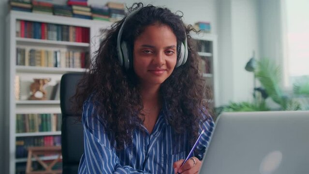 Young beautiful black haired Indian woman student in wireless headphones looks at camera and holds up pencil preparing thesis sits at desk in home office. Education, training, technology