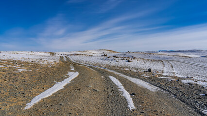 A dirt road winds through a snow-covered hilly valley. Rocks are scattered around. Blue sky with picturesque radial clouds. Altai © Вера 