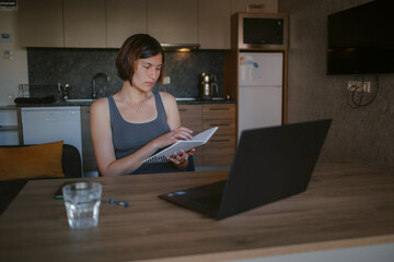 asian female freelancer working on laptop in living room. Focused lady studying distantly and making notes in copybook, listening to lecture. technologies for online education and self classes