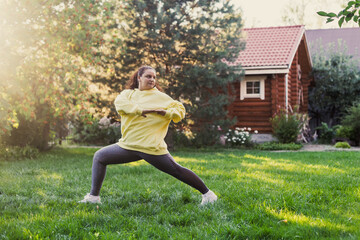 Middle-aged, female adult doing physical exercises stretching to keep fit wearing sports clothes on backyard on green grass with wooden country house and tall trees in background. Body positive