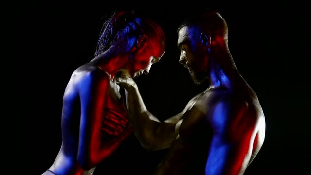 a couple with golden metallic skin stands in profile on a black background. a man strokes a woman's shoulder. red, blue and white light. the dark key. the middle plan