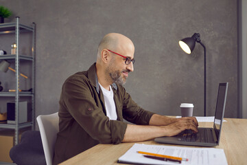 Smiling middle-aged man in spectacles work online on computer consult client in modern home office...