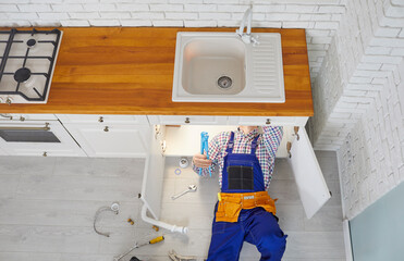 Plumber working in a modern kitchen. Plumber in an overall uniform lying down on the floor under the sink drain, repairing pipes or changing a rubber gasket. Top view, overhead, from above, high angle