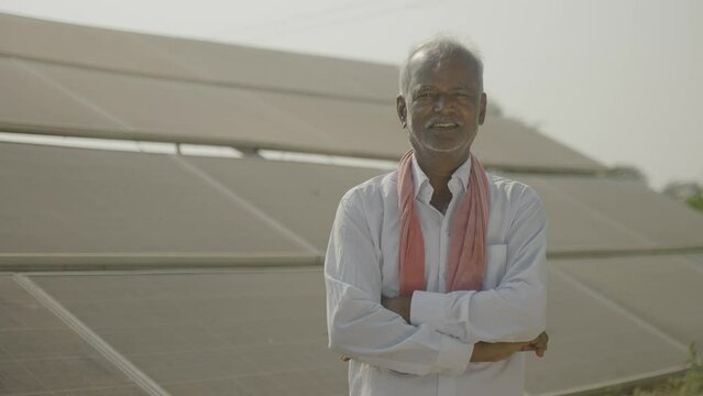 handheld shot of Smiling village farmer standing with crossed arms by looking at camera in front of solar panel - concept of modern agriculture, green energy and eco-friendly farming.