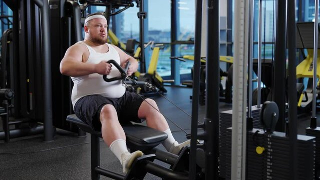 Fat burning, overweight funny man lifting blocks on rowing machine, training on block device and gym equipment.