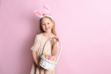 Obraz na płótnie Canvas Happy Easter. A cute beautiful girl with bunny ears holds a basket of color eggs in her hands. Pink background. Space for text