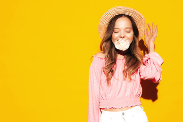 Closeup portrait of young beautiful smiling female in trendy summer  clothes. Sexy carefree woman posing near yellow wall in studio. Positive model holding flower in her mouth. In hat