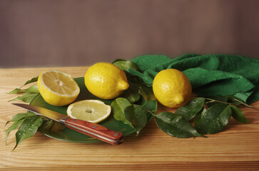 Three lemons with leaves, a plate and a knife on a table with a wooden texture. 