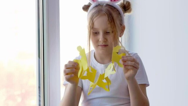 A petite blonde with freckles on her face and bunny ears unrolls and reveals a garland of bunnies cut out of yellow paper. Easter concept for DIY home decor.