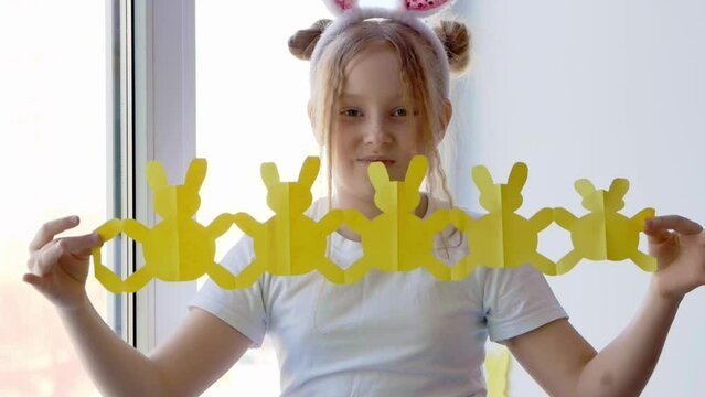 A petite blonde with freckles on her face and bunny ears unrolls and reveals a garland of bunnies cut out of yellow paper. Easter concept for DIY home decor.