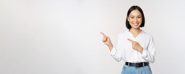 Image of smiling young office lady, asian business entrepreneur pointing fingers left, showing client info, chart of banner aside on copy space, white background - obrazy, fototapety, plakaty