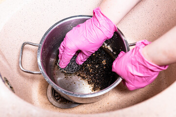 above view of gloved hands wash pan with burnt food with metal sponge in pink sink at home kitchen