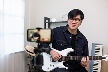 Young asian musician teaching the rock guitar class online live streaming on smartphone with student. Korean man playing electric guitar singing a song and record by cell phone shaed online social