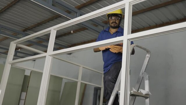 worker fixing screw using drilling machine or gun on aluminium partition frame at construction site - concept of maintenance service, blue collar jobs and safety.
