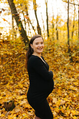 Beautiful pregnant woman in a black dress in the autumn park. Golden autumn. Fallen leaves. Awaiting the birth of a child.