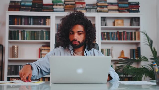 Young focused curly Arabian man student making notes in workbook for online lesson typing on laptop keyboard studying at internet college sits at table in front of bookcase. Distance education concept