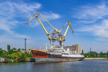 Obraz premium Mykolaiv, Ukraine - July 26, 2020: A large ship against the background of two port cranes on the Inhul River in Mykolaiv. View from the pedestrian bridge to the old rusty abandoned vessel