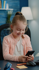 Young girl using smartphone with touch screen at desk. Little child smiling while looking at phone screen after class lesson and school education. Schoolgirl having fun on modern device