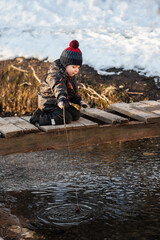Naklejka premium A little boy in the park in early spring. A child is playing with a homemade fishing rod made of a stick. A carefree childhood.