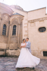 Stylish groom in a suit and a beautiful bride in a white dress are hugging on the background of the building. Wedding portrait of newlyweds in love.