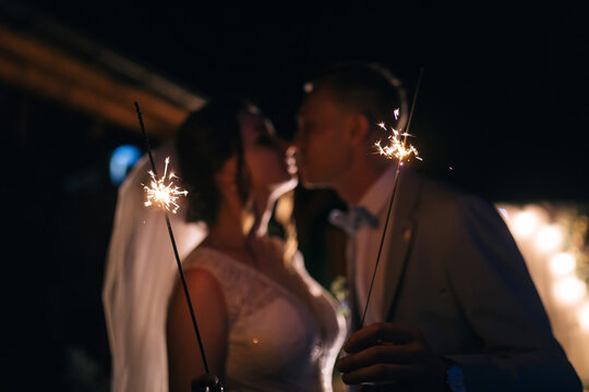 newlyweds with sparklers kiss and say goodbye to guests in a night restaurant. beautiful end of the wedding day. The groom in a suit and the bride in a white elegant dress.