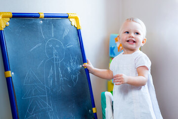 cute little female artist paints on an easel with chalk while standing on a chair.