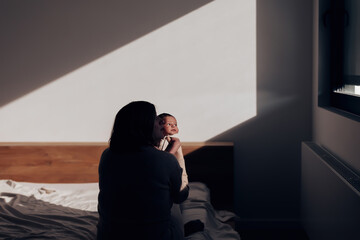 chiaroscuro portrait of a newborn baby - infant held in her mother's arms on the bed looking out the window - light and shadow