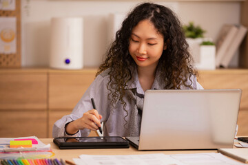 In the morning, an Asian designer works on a tablet with a stylus, sketching and writing designs.