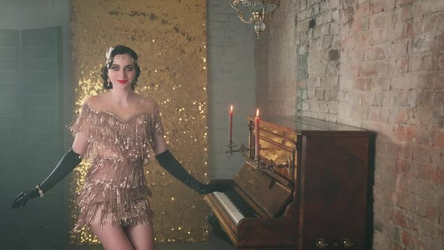 Happy beautiful young woman in bright sparkling luxury gold dress, smiles, gown fluttering, dances in room near piano, candles burn. Girl cheerful joyful dancer in motion. 20s stylized carnival party.