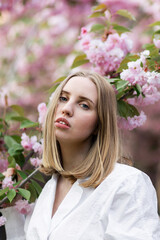 Fototapeta premium A blonde woman in a white shirt poses near the cherry blossoms. Spring mood