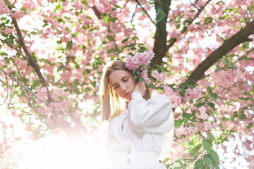 A blonde woman in a white shirt poses near the cherry blossoms. Spring mood