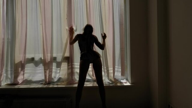 The silhouette of a sexy woman in underwear and striptease shoes who dances a sensual dance near a large window with long curtains.
