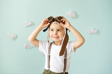 A boy in an aviator helmet and suspenders stands on a blue background with clouds and smiles.