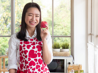 Pretty young Asian woman wearing red heart shape pattern apron holding red apple in the kitchen at home.