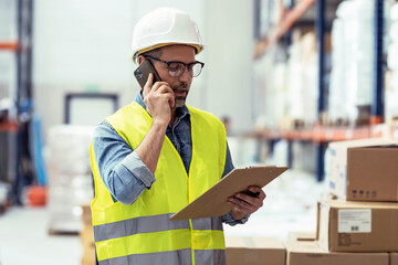 Mature worker talking with smartphone while passing list of material that has arrived at the store warehouse.