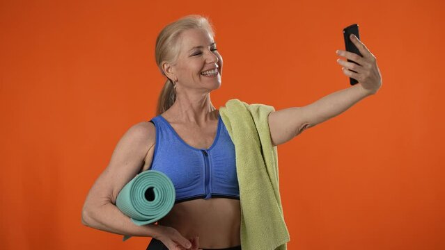 Smiling mature strong sporty athletic fitness woman taking selfie with smart phone, isolated on plain orange background