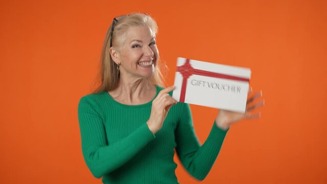 Happy mature blonde woman 40s 50s dancing with gift certificate voucher winner gesture looking camera isolated on orange color background studio portrait.