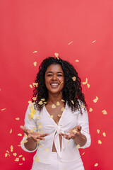 Mid waist portrait of cheerful woman playing with party confetti in red background. Vertical front view of african american woman on festive celebration with confetti. People background concept.