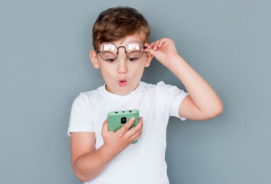 Shocked child boy using smartphone and taking off glasses on gray background