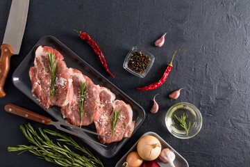 three pieces of pork neck with rosemary, pepper, onion, garlic on a dark tray, a knife and a fork on a black background for cooking. top view. - Powered by Adobe
